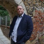 Phil Creighton leaning against an archway that was once part of Reading Abbey. There is red brickwork behind the arch. On the left there is a footpathy and trees and metal fencing can be seen behind it.