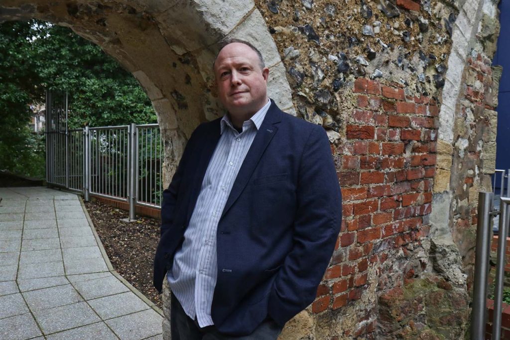 Phil Creighton leaning against an archway that was once part of Reading Abbey. There is red brickwork behind the arch. On the left there is a footpathy and trees and metal fencing can be seen behind it.