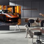 A man watching an LG television in a lounge with a Dalmatian next to him. The man is standing up and has a remote control in his hands. In the backrgound there is a kitchen. The television is massive and has a flash car on the screen.