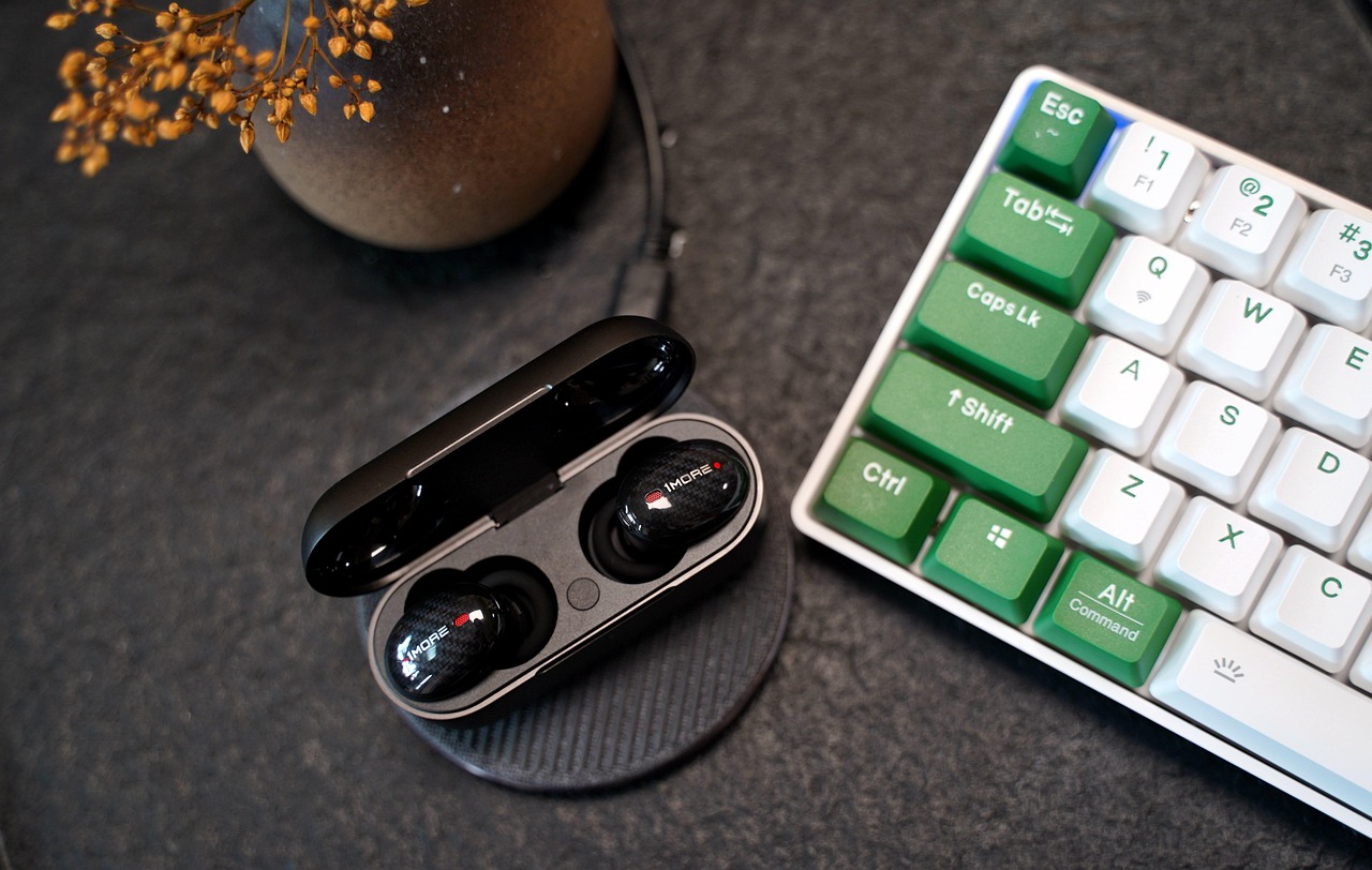 A pair of Bluetooth earbuds in their case next to a keyboard. Some of the command keys are in green. In the top left hand corner is a plant pot.