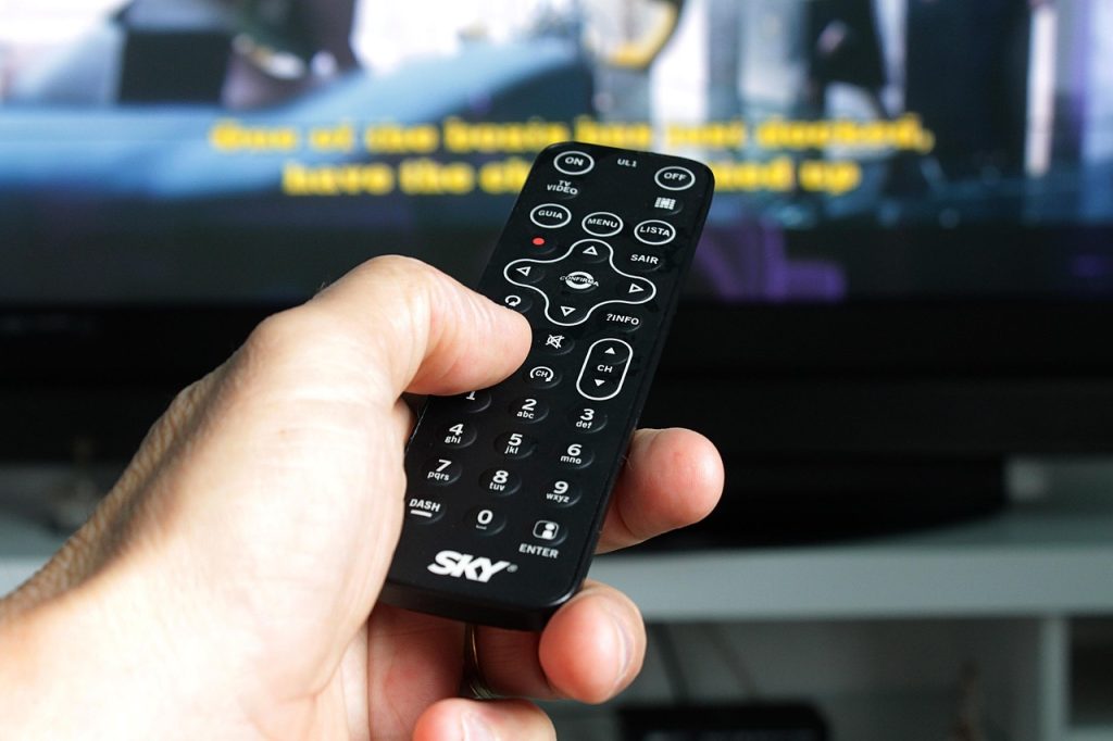 A remote control in a hand with a thumb about to press a button. A television screen is in the background. Auracast