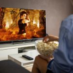 A person watching television. They are wearing a blue shirt and we are seeing their back. On their lap is a bowl of popcorn. The screen features an action movie scene of a woman walking through an explosion in a street. Auracast televisions are on the market but not always on show