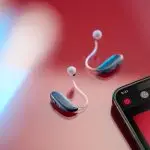 The ReSound Vivia hearing aids on a red background. On the right hand side of the image is a mobile phone showing the ReSound app