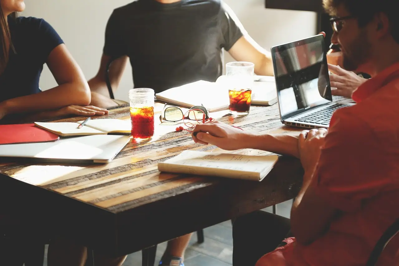 People sitting around a table at a business meeting. We cannot see faces, only torsos and arms. There are drinks, paperework and a laptop on the table. The picture helps illustrate a meeting where Auracast Advocates are sharing information about the Bluetooth technology