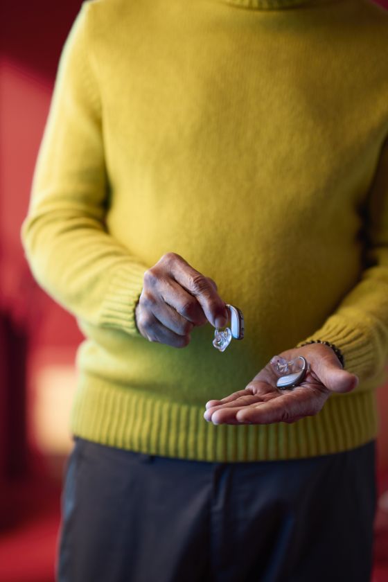A person holding a ReSound Enzo IA  hearing aid in each hand. They are wearing a yellow jumper