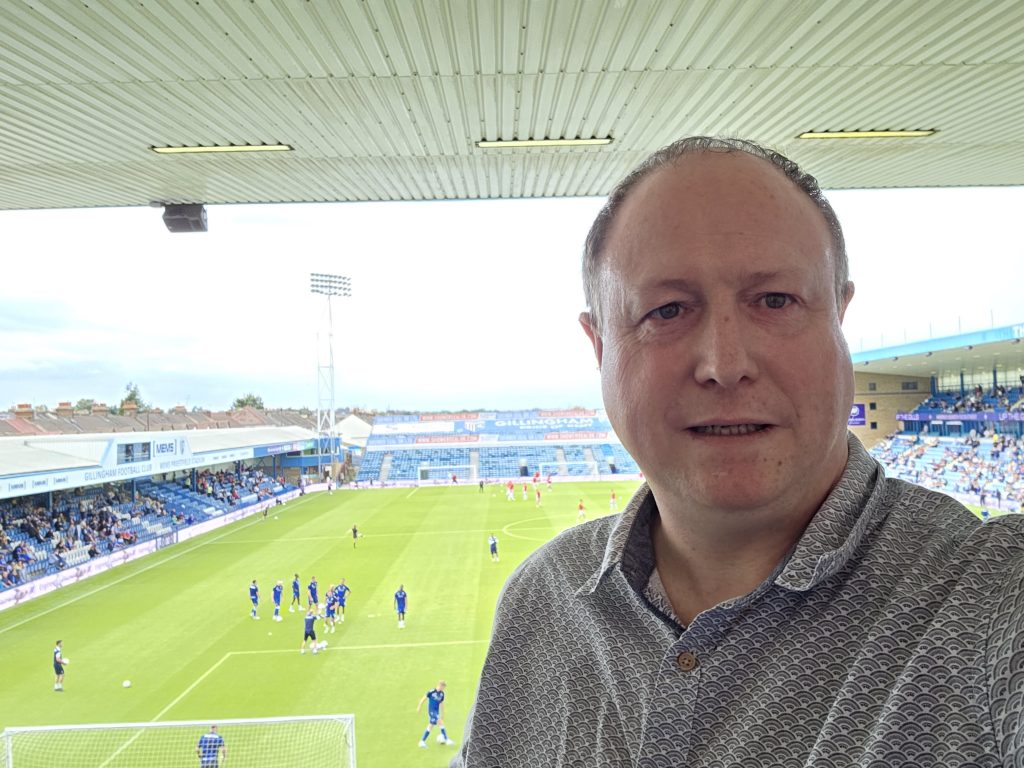 Phil Creighton on the right with a football stadium behind him. The roof of the stand can be seen behind him with a speaker attached to it 