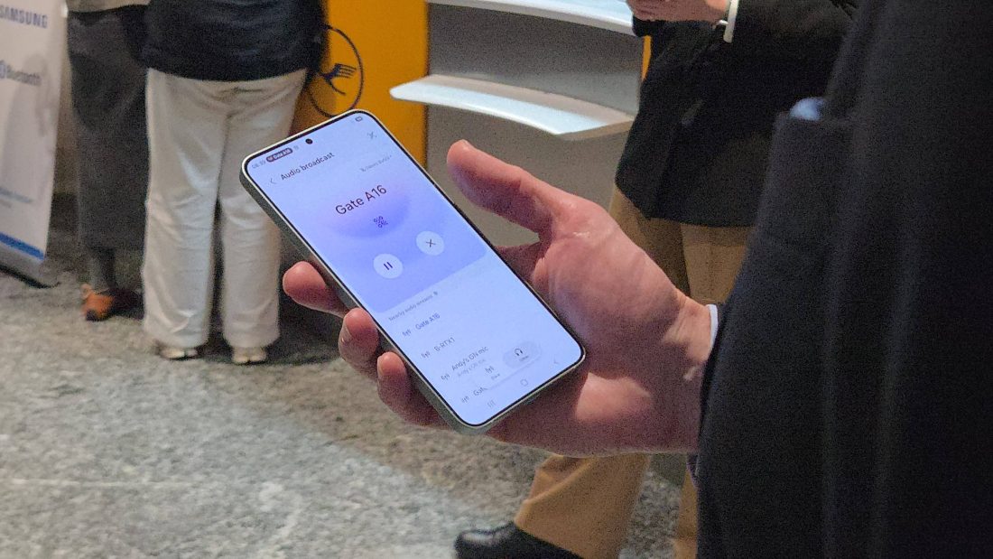 A person holding a mobile phone at Frankfurt Airport showing they are tuned into the Auracast at Gate A16