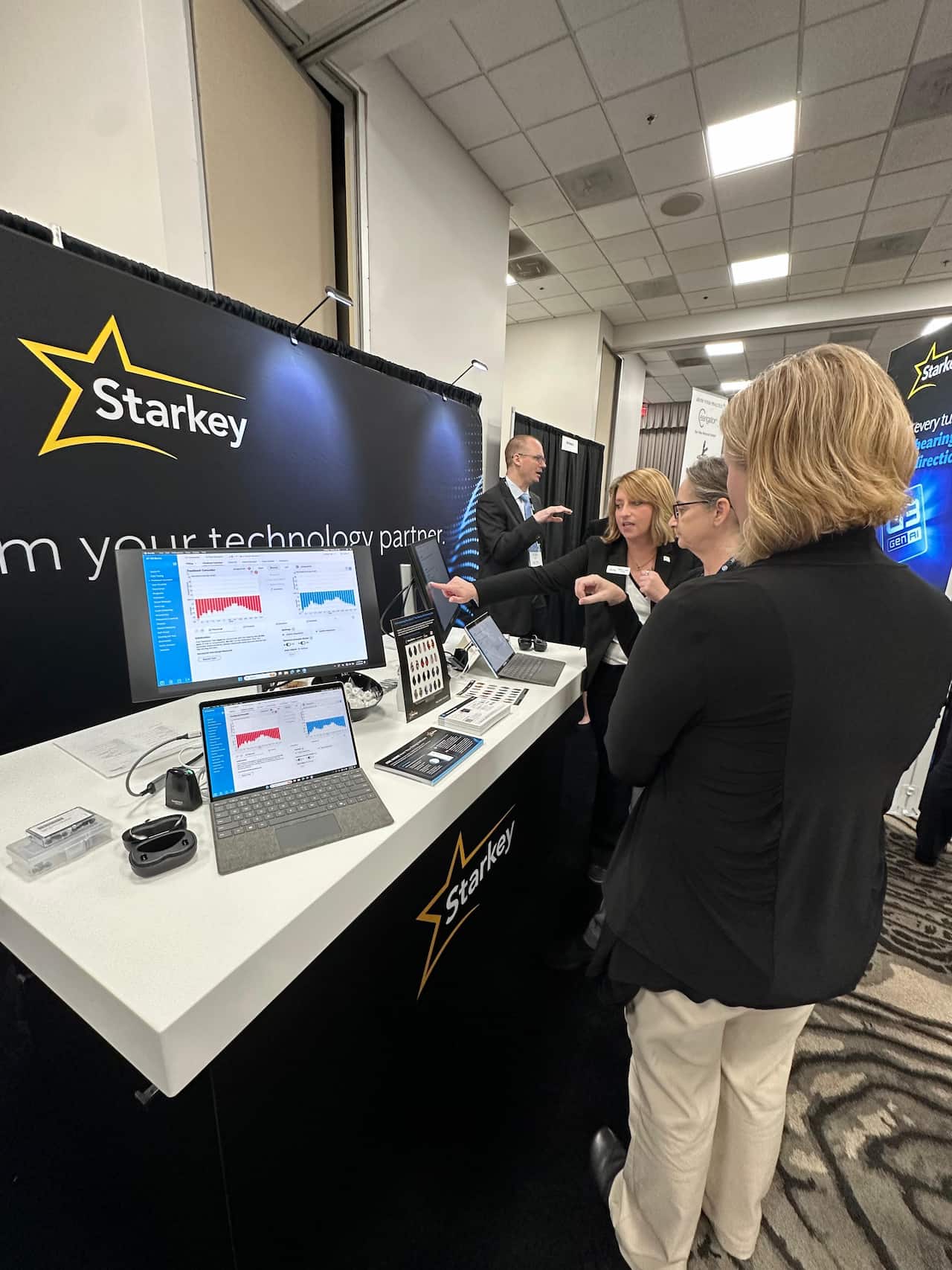 A woman in a black jacket points at a computer screen displaying data charts at a Starkey exhibit booth, while three other individuals observe and discuss the technology. The booth features professional signage with the Starkey logo and the phrase "I am your technology partner." Laptop computers and informational materials are arranged on the white countertop. Auracast