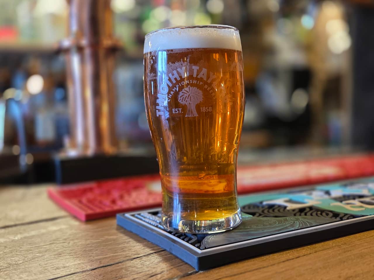 A close-up, shallow depth-of-field photo of a pint of amber ale in a Timothy Taylor glass sitting on a bar mat in a pub