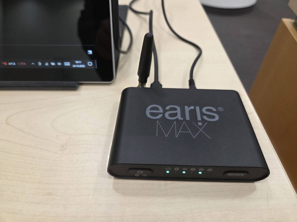 The EarisMax Auracast transmitter on a desk with lights on. Cables are coming from its rear and attached to a tablet, the screen of which we can just see in the top left