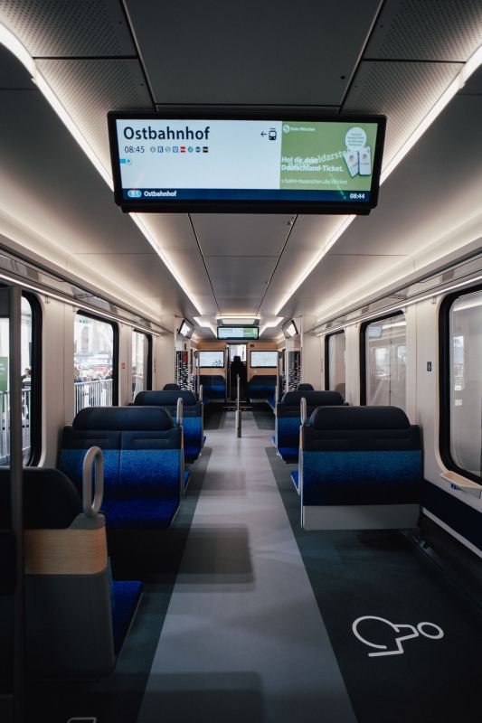 View down the center aisle of a modern train carriage with blue seats and large digital information screens hanging from the ceiling