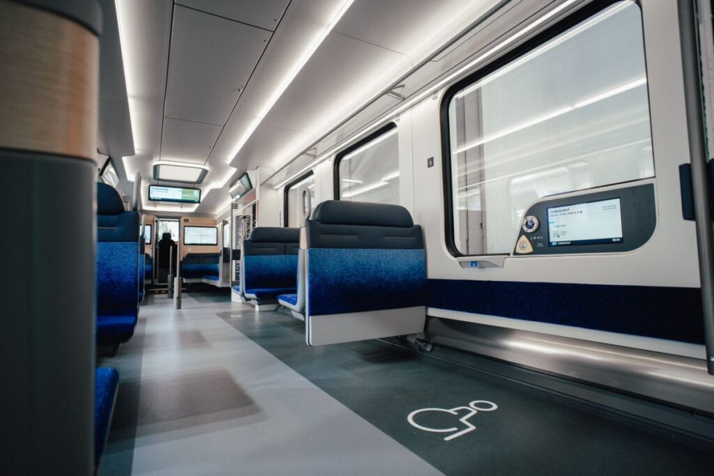 Interior of a train showing a designated wheelchair space on the floor and a digital screen near the window displaying station information