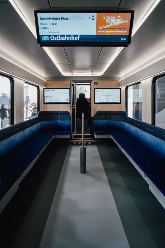 View down the center aisle of a modern train carriage with blue seats and large digital information screens hanging from the ceiling