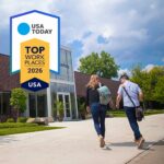 Starkey is celebrating its USA Today Top Workplaces award. The image shows the award logo and two people walking towards the entrance of Starkey HQ. They have their backs to us and we can see one has a rucksack, the other a satchel. It's a bright sunny day with blue sky and happy clouds