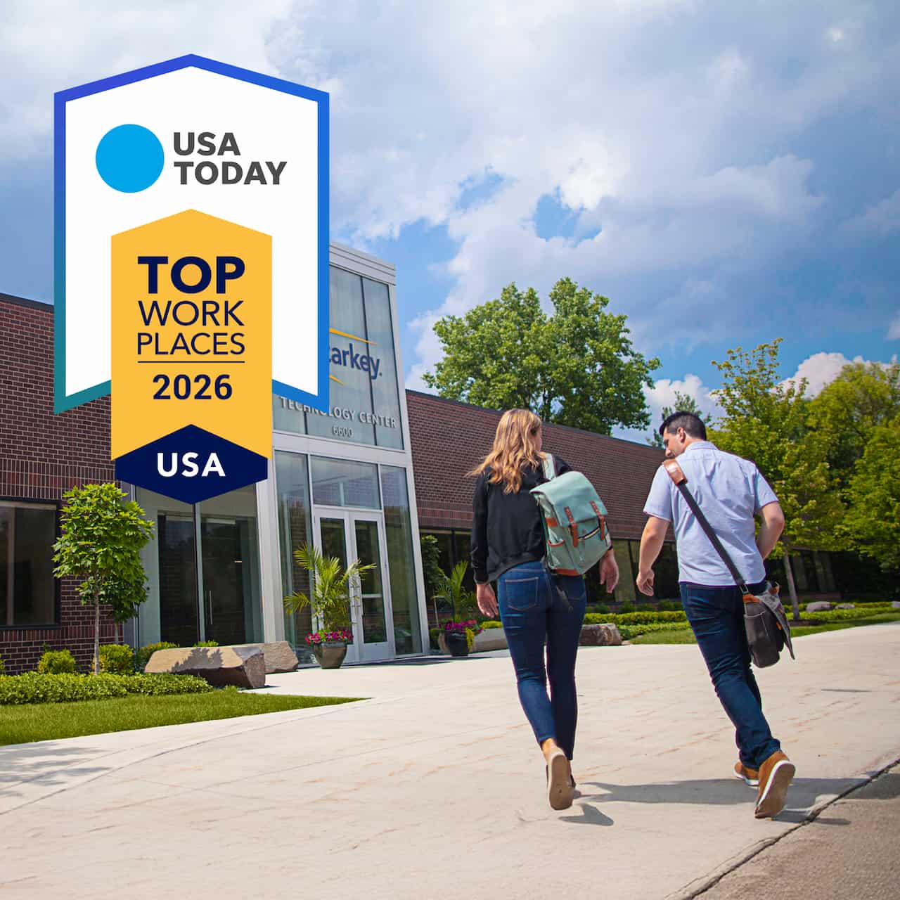 Starkey is celebrating its USA Today Top Workplaces award. The image shows the award logo and two people walking towards the entrance of Starkey HQ. They have their backs to us and we can see one has a rucksack, the other a satchel. It's a bright sunny day with blue sky and happy clouds