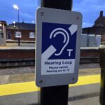 A close-up photograph of an official accessibility sign mounted on a black post at an outdoor railway platform during dusk. The rectangular grey sign features a prominent blue and white international symbol for hearing accessibility, showing a stylized ear shape crossed by a diagonal line next to a capital letter T. Below the icon, the text reads Hearing Loop, followed by the instruction Please switch hearing aid to T. The background shows a yellow tactile paving strip along the platform edge, train tracks, and a brick station building under a cloudy evening sky with bright station lights illuminated.