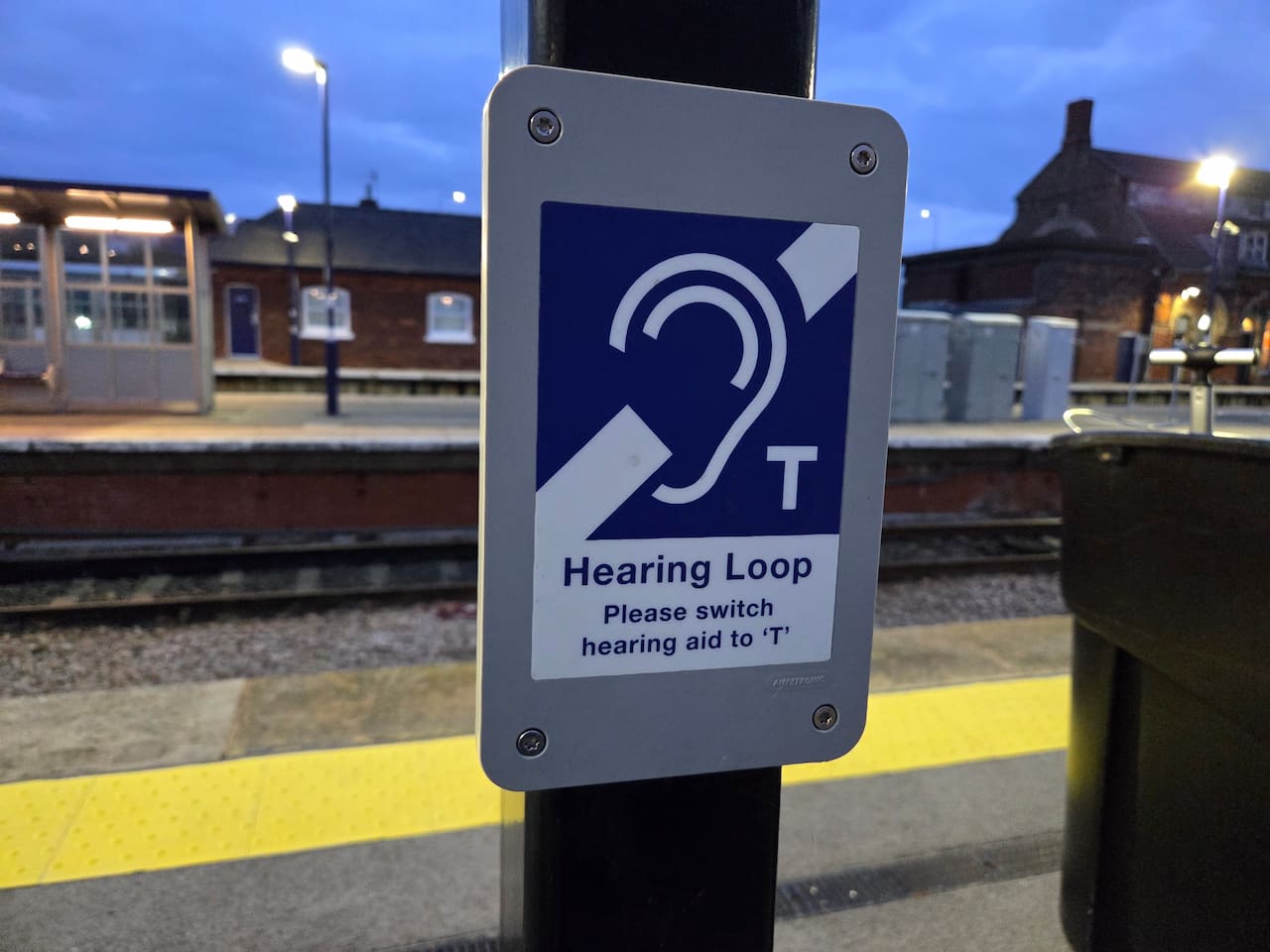 A close-up photograph of an official accessibility sign mounted on a black post at an outdoor railway platform during dusk. The rectangular grey sign features a prominent blue and white international symbol for hearing accessibility, showing a stylized ear shape crossed by a diagonal line next to a capital letter T. Below the icon, the text reads Hearing Loop, followed by the instruction Please switch hearing aid to T. The background shows a yellow tactile paving strip along the platform edge, train tracks, and a brick station building under a cloudy evening sky with bright station lights illuminated.