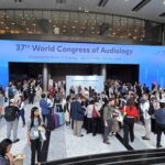 A high-angle, wide shot of a bustling convention center lobby during the 37th World Congress of Audiology. Above the main entrance to the Grand Ballroom, a massive digital screen spans the wall, featuring a bright blue gradient background with the text "37th World Congress of Audiology" and the theme "Broadening the Horizon of Audiology". Below, a diverse crowd of international delegates in business casual attire stands around tables, networking and talking on the polished marble floor.