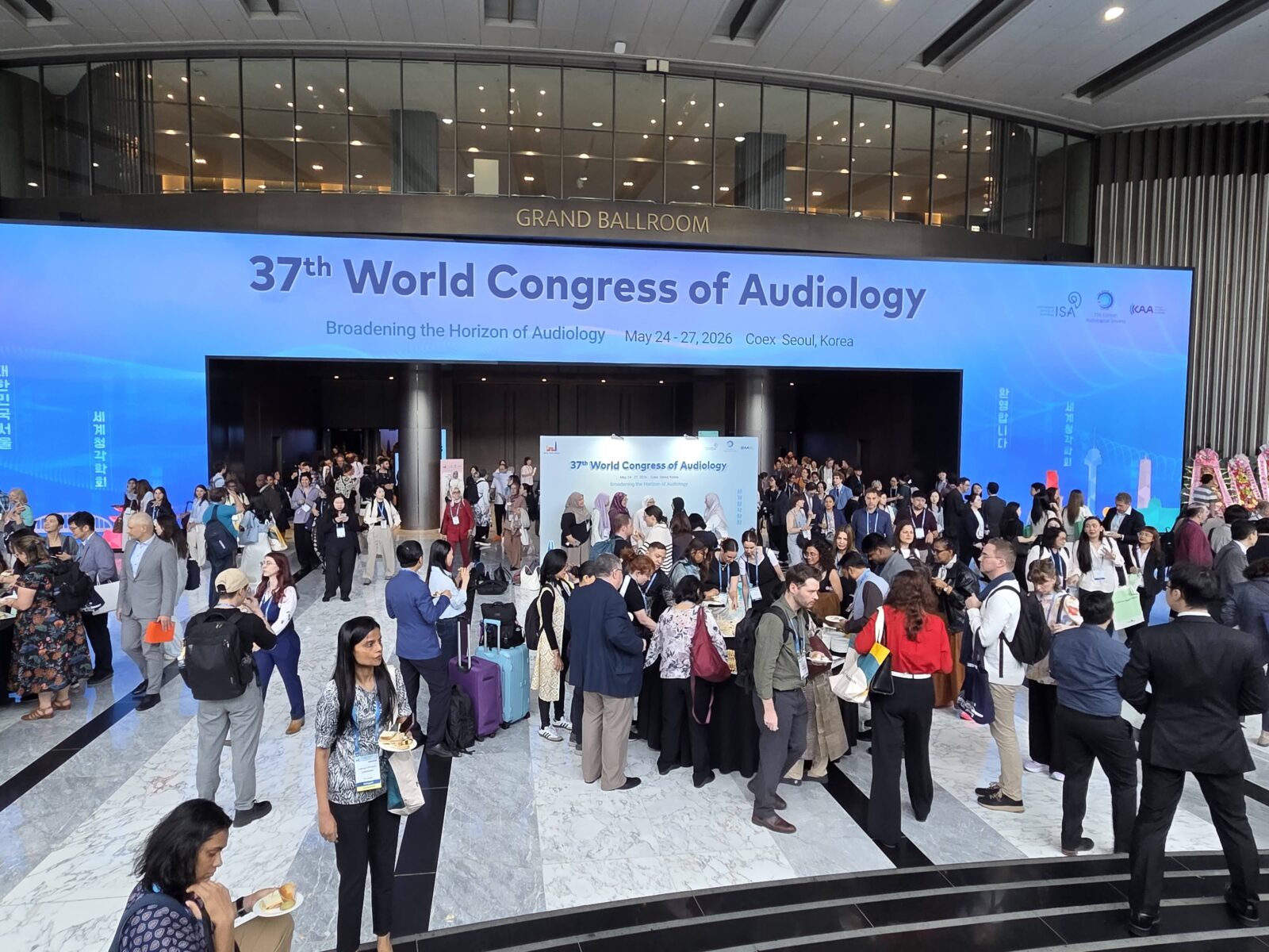 A high-angle, wide shot of a bustling convention center lobby during the 37th World Congress of Audiology. Above the main entrance to the Grand Ballroom, a massive digital screen spans the wall, featuring a bright blue gradient background with the text "37th World Congress of Audiology" and the theme "Broadening the Horizon of Audiology". Below, a diverse crowd of international delegates in business casual attire stands around tables, networking and talking on the polished marble floor.