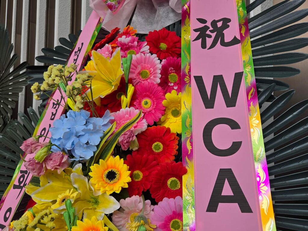 A vibrant, close-up shot of a traditional congratulatory flower wreath at the convention. The arrangement is densely packed with colorful blossoms, including bright pink, red, and yellow gerbera daisies, yellow lilies, and blue hydrangeas. Flanking the flowers are wide vertical ribbons; the prominent pink ribbon on the right features a large black Hanja character at the top with the letters "WCA" printed vertically below it.