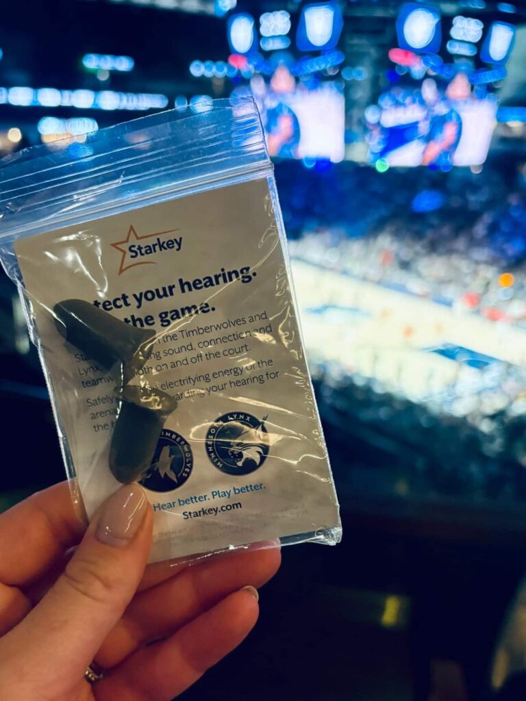 A close-up shot of a hand holding a small transparent plastic packet containing a pair of dark grey foam earplugs. The packet features the Starkey logo alongside the Minnesota Lynx and Minnesota Timberwolves logos. Text on the packaging reads "Protect your hearing. Love the game." and "Hear better. Play better. Starkey.com". The background shows a blurry, brightly lit basketball arena during a game.