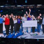 A group of 11 people standing on a basketball court, smiling and waving at the camera. Two women in the center hold a large, oversized ceremonial cheque made out to Girls Taking Action for the amount of eighty thousand dollars, dated May 27, 2026. The Minnesota Lynx logo is visible on the court floor underneath them, and a crowded arena stands in the background.