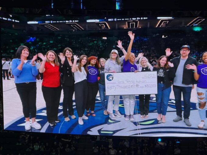 A group of 11 people standing on a basketball court, smiling and waving at the camera. Two women in the center hold a large, oversized ceremonial cheque made out to Girls Taking Action for the amount of eighty thousand dollars, dated May 27, 2026. The Minnesota Lynx logo is visible on the court floor underneath them, and a crowded arena stands in the background.