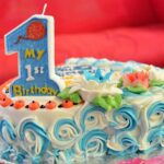 A close-up photograph of a decorated first birthday cake on a silver foil tray. The cake features white frosting with intricate blue and white piped rosettes along the sides and delicate floral icing decorations on top, including a white lotus flower with a blue center and a coral-colored flower. On the left side, a large, upright, glittery blue candle in the shape of the number one reads My 1st Birthday with a small orange balloon graphic at the top. The edge of the cake is dotted with small orange icing details, and a red sofa is softly blurred in the background.