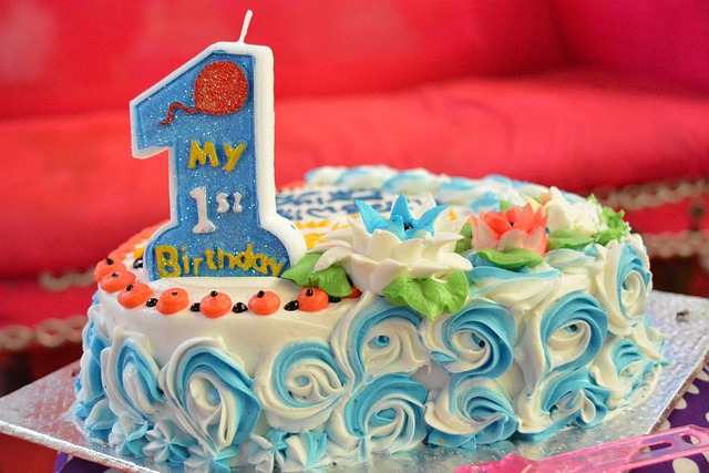 A close-up photograph of a decorated first birthday cake on a silver foil tray. The cake features white frosting with intricate blue and white piped rosettes along the sides and delicate floral icing decorations on top, including a white lotus flower with a blue center and a coral-colored flower. On the left side, a large, upright, glittery blue candle in the shape of the number one reads My 1st Birthday with a small orange balloon graphic at the top. The edge of the cake is dotted with small orange icing details, and a red sofa is softly blurred in the background.