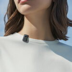 A close-up shot focusing on the collar of a person wearing a plain white t-shirt. Clipped onto the neckline is a tiny, rectangular black wireless microphone, the Emendion Aspen Aura. The compact 5g device features a small green indicator light on the side and subtle branding on the front casing, demonstrating its miniature size and portability against a clear blue sky background.
