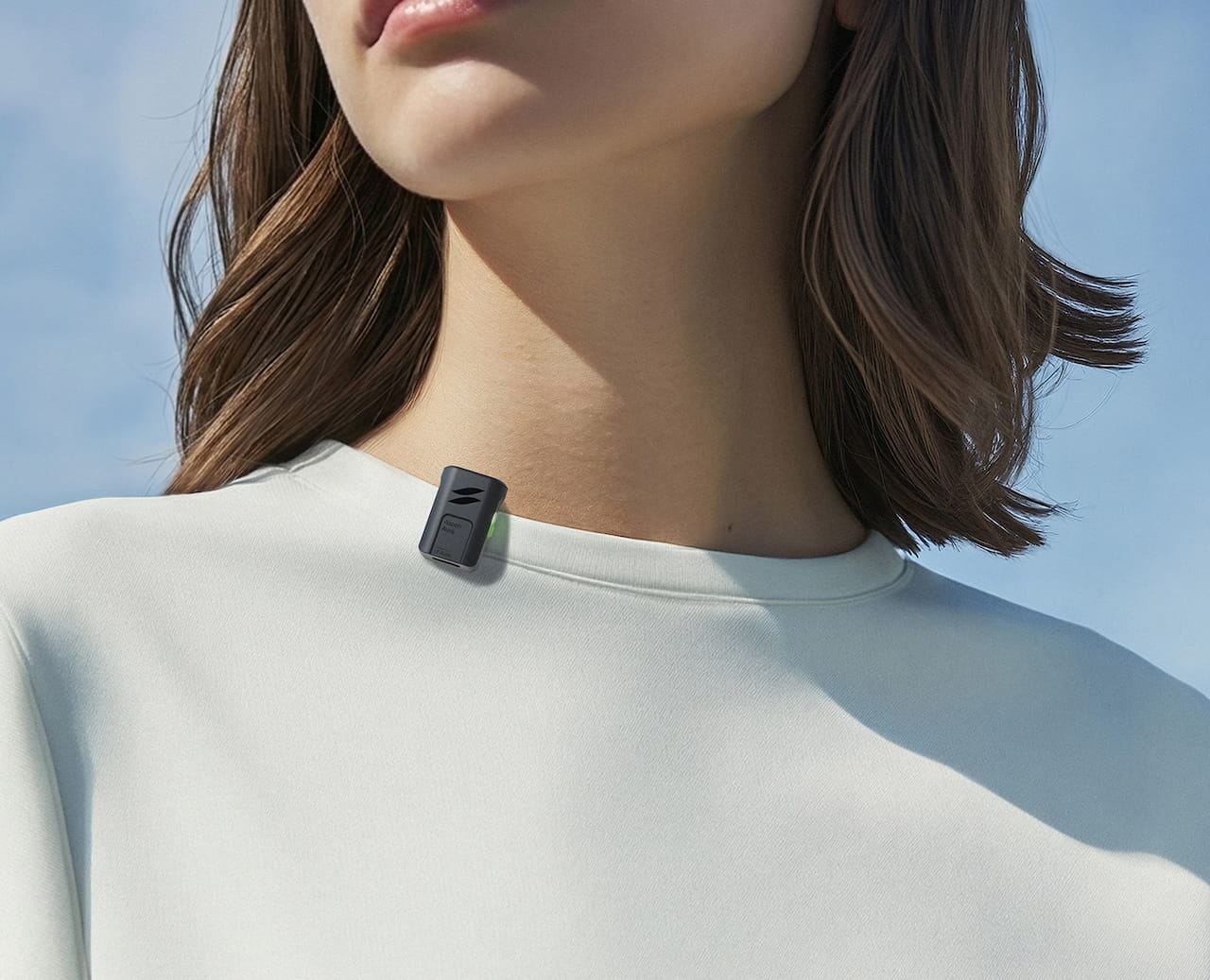 A close-up shot focusing on the collar of a person wearing a plain white t-shirt. Clipped onto the neckline is a tiny, rectangular black wireless microphone, the Emendion Aspen Aura. The compact 5g device features a small green indicator light on the side and subtle branding on the front casing, demonstrating its miniature size and portability against a clear blue sky background.