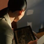 A close-up photograph taken from a high angle behind a person with dark hair tied back in a low ponytail, showcasing a discreet modern hearing aid resting over the top of their right ear. The individual is wearing a light sage green tailored blazer and is looking down, holding a framed piece of artwork in a softly lit room with dramatic shadows cast on the wall in front of them. The image illustrates the real-world application of the advanced hearing care technology behind the rising global sales trends
