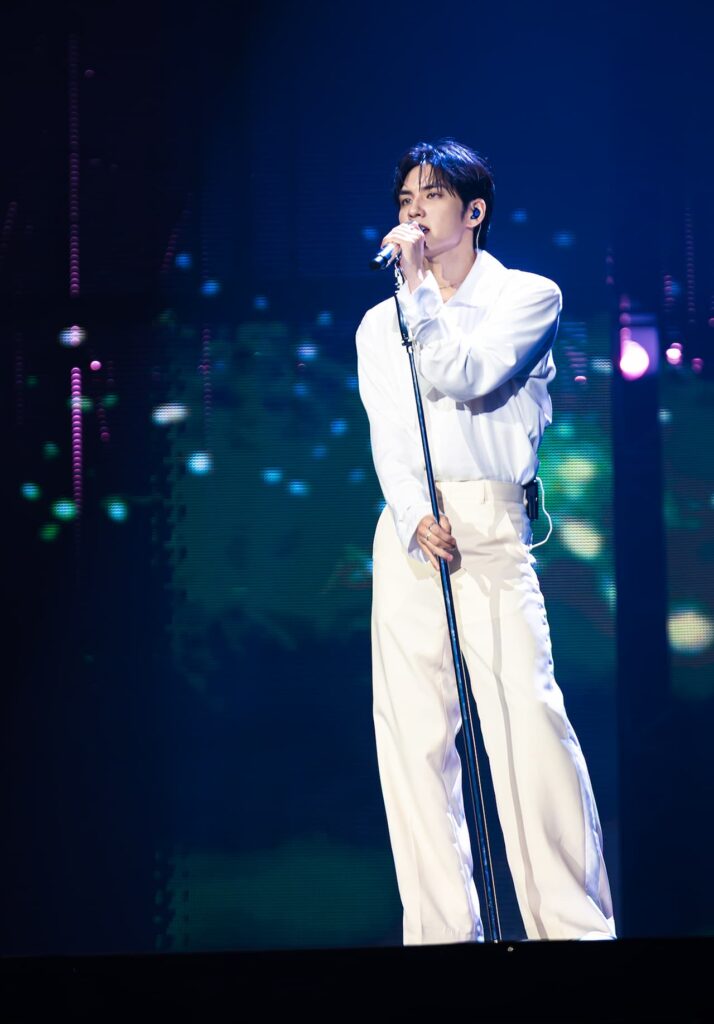 A full-length, eye-level shot of South Korean singer WONPIL performing live on stage at his solo concert, Unpiltered. He is wearing a long-sleeved white button-down shirt tucked into loose-fitting cream trousers. He holds a silver microphone close to his mouth with both hands while standing behind a microphone stand. The background is a dark, vibrant stage screen filled with soft, out-of-focus bokeh lights in shades of blue, green, and pink.