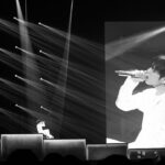 A wide, dramatic black and white photograph of the Unpiltered concert stage. On the left side of the stage, the artist sits on a raised platform, illuminated by a grid of multiple sharp spotlights casting geometric beams of light across the darkness. On the right side of the stage, a massive, towering LED screen shows a live, high-contrast close-up broadcast of the artist singing passionately into his microphone.