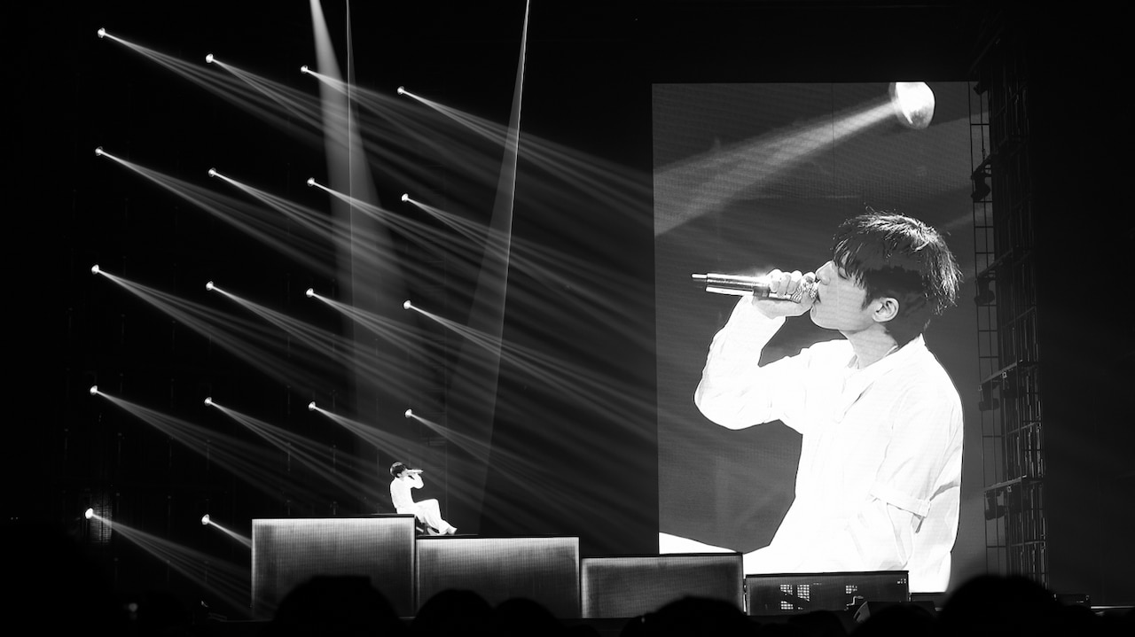 A wide, dramatic black and white photograph of the Unpiltered concert stage. On the left side of the stage, the artist sits on a raised platform, illuminated by a grid of multiple sharp spotlights casting geometric beams of light across the darkness. On the right side of the stage, a massive, towering LED screen shows a live, high-contrast close-up broadcast of the artist singing passionately into his microphone.