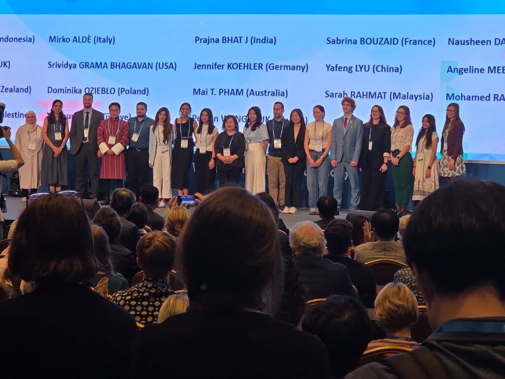 A wide shot from the back of an auditorium showing a large group of international delegates standing in a long row on a brightly lit stage. Behind them, a massive digital screen displays names and corresponding countries in black text against a light blue background, including visible entries like "Mirko ALDÈ (Italy)", "Prajna BHAT J (India)", "Sabrina BOUZAID (France)", and "Jennifer KOEHLER (Germany)". The delegates are dressed in professional and traditional attire, looking toward the camera. In the foreground, the back of the heads of several audience members are visible as they sit in darkened rows watching the presentation.