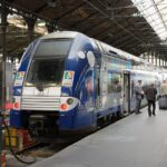 A double decker SNCF train with a blue and silver front sits stationary at a platform under the large iron and glass arched roof of Gare Saint Lazare station in Paris. The train features the Region Haute Normandie logo on the front livery. Several passengers walk along the concrete platform next to the train, including a man pulling a suitcase, while a railway worker in uniform stands near one of the open carriage doors.