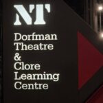 An illuminated dark grey directional sign at night outside the National Theatre in London. The sign features the white glowing NT logo at the top, followed by the words Dorfman Theatre & Clore Learning Centre in a white serif font, with a large red arrow pointing right.