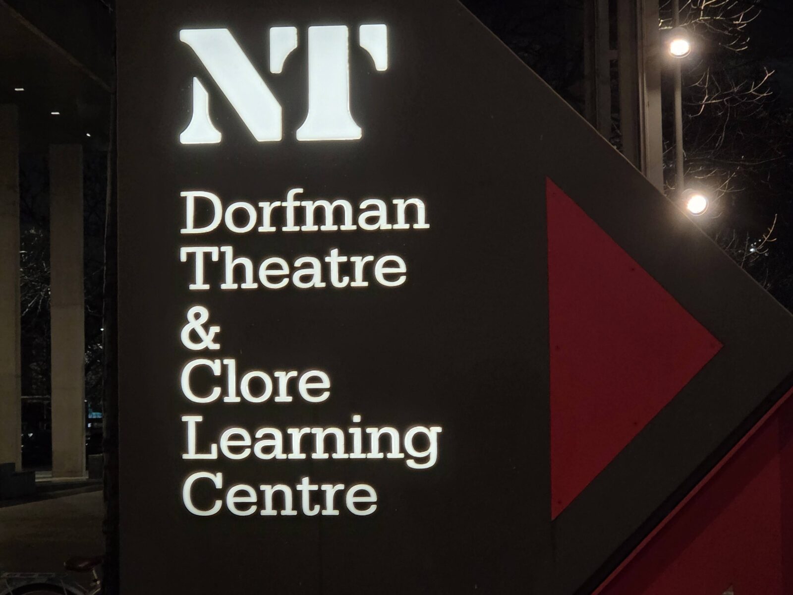 An illuminated dark grey directional sign at night outside the National Theatre in London. The sign features the white glowing NT logo at the top, followed by the words Dorfman Theatre & Clore Learning Centre in a white serif font, with a large red arrow pointing right.