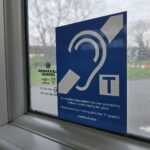 A blue and white sticker affixed to the inside of a window, showing the international symbol for deafness—a stylized ear outline with a diagonal line through it. Next to the ear is a square icon with a capital letter T. Below the graphics, text reads: An induction loop system has been provided by Ardent to assist hearing aid users. Please switch your hearing aid to the "T" position. ardent-uk.com. Out of focus trees and a road are visible through the glass.