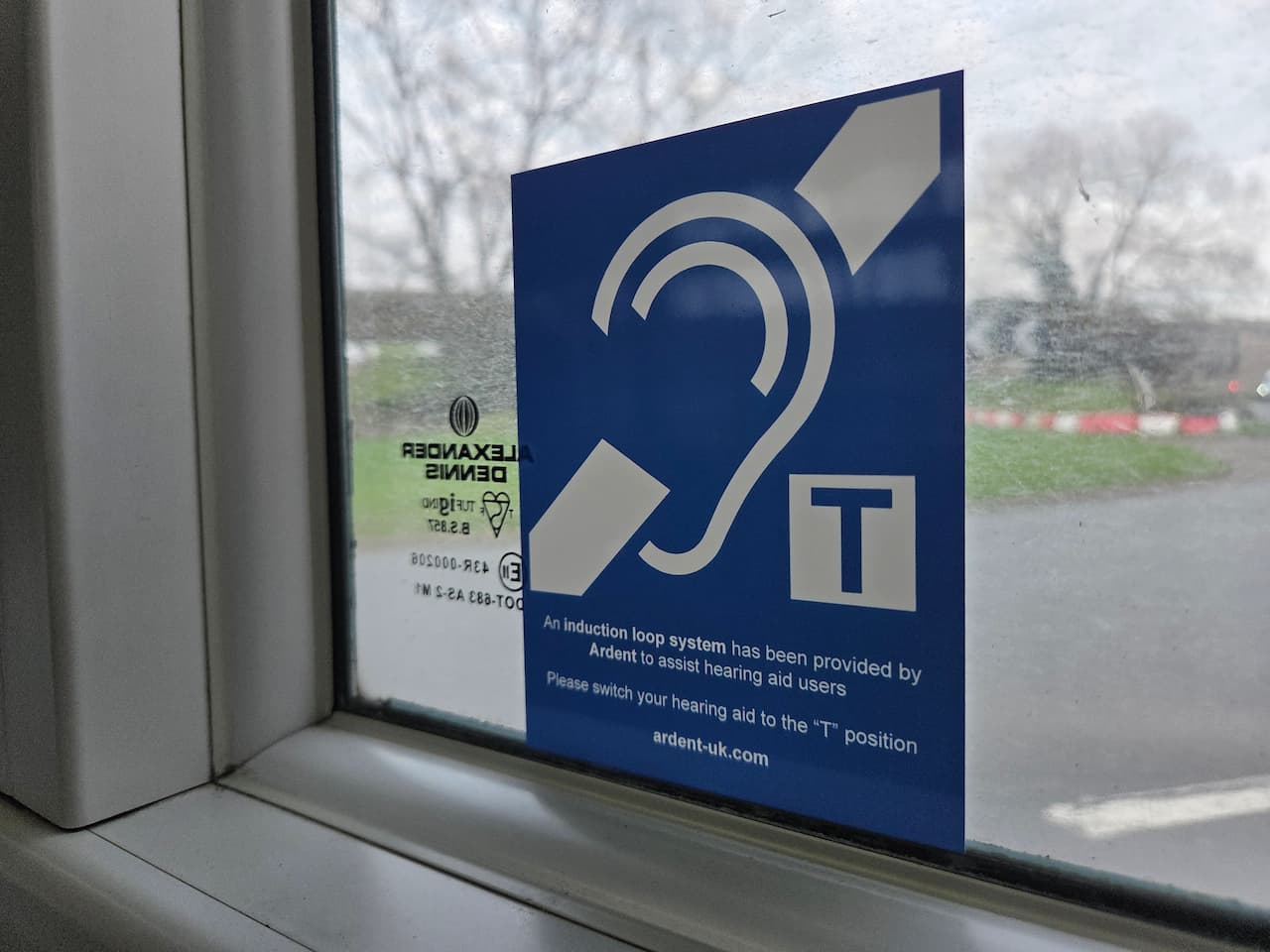 A blue and white sticker affixed to the inside of a window, showing the international symbol for deafness—a stylized ear outline with a diagonal line through it. Next to the ear is a square icon with a capital letter T. Below the graphics, text reads: An induction loop system has been provided by Ardent to assist hearing aid users. Please switch your hearing aid to the "T" position. ardent-uk.com. Out of focus trees and a road are visible through the glass.