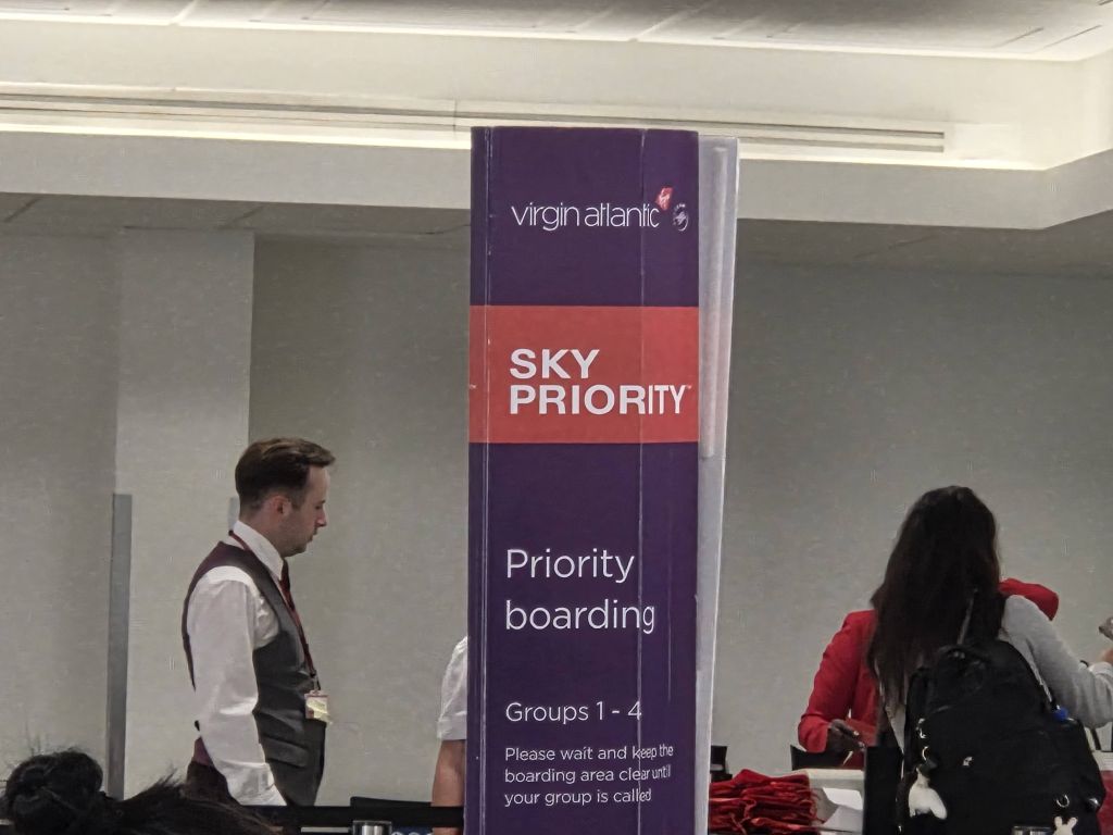 A tall purple and red column sign at an airport gate lounge that reads Virgin Atlantic, Sky Priority, Priority boarding, Groups 1-4, and instructions to keep the area clear. An airport gate agent is visible on the left, and passengers are gathering on the right.