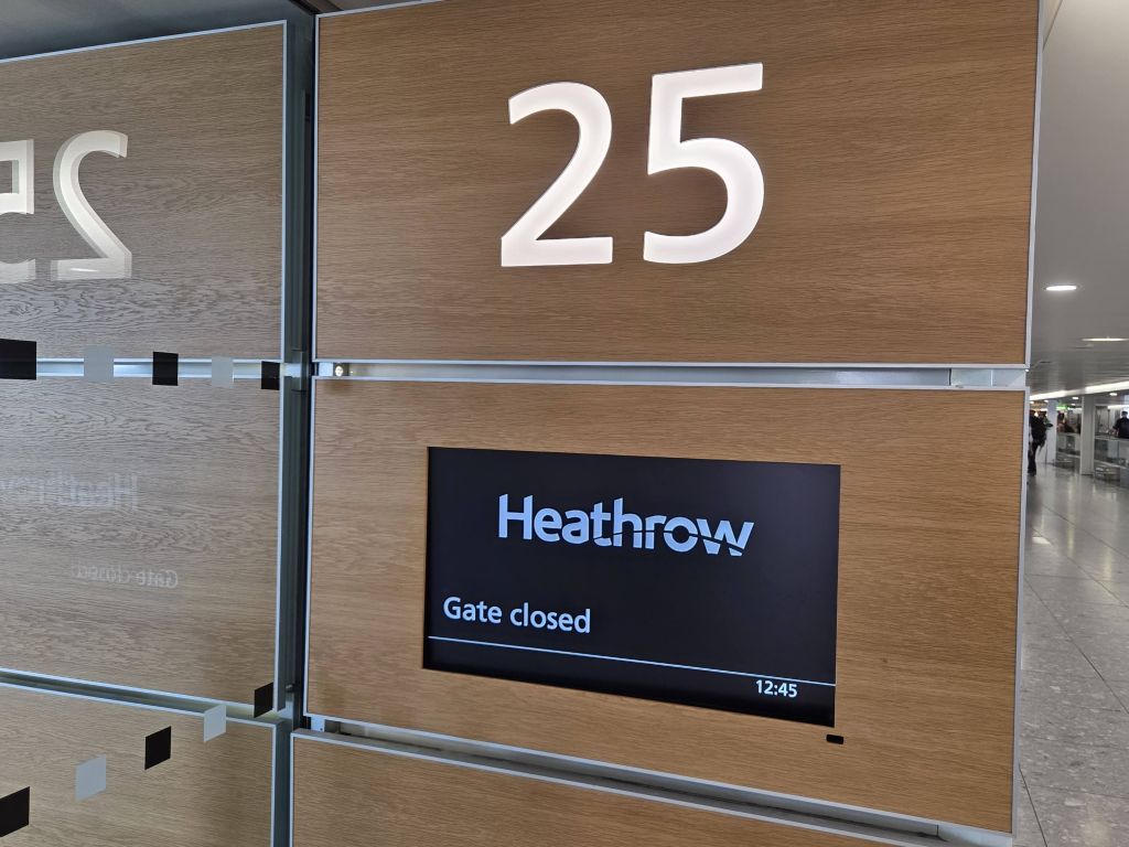 A close up shot of airport gate signage with a wood grain background panel. The white glowing text at the top reads 25. Below it is an integrated digital screen showing a black background with the white Heathrow logo, the text Gate closed, and a digital clock reading 12:45.