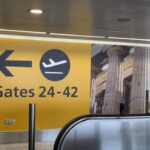 An airport terminal wall painted bright yellow with a black left pointing arrow, an airplane symbol, and the text Gates 24-42. A large graphic wallpaper of ancient Greek pillars is to the right, and the edge of a silver metal escalator handrail is visible in the foreground.