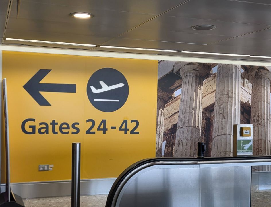 An airport terminal wall painted bright yellow with a black left pointing arrow, an airplane symbol, and the text Gates 24-42. A large graphic wallpaper of ancient Greek pillars is to the right, and the edge of a silver metal escalator handrail is visible in the foreground.