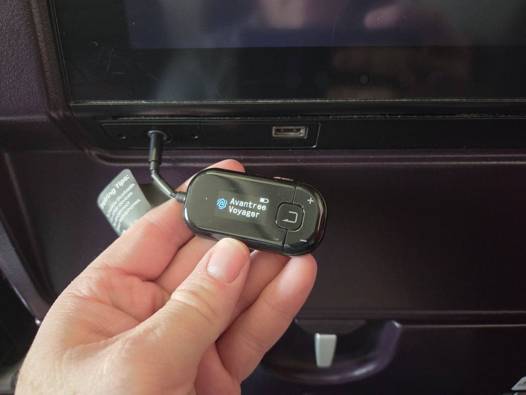 A hand holding a small black electronic transmitter called an Avantree Voyager in front of an airplane seatback entertainment screen. The transmitter screen is illuminated showing its name, and a small black cable plugs it directly into the screen headphone jack.