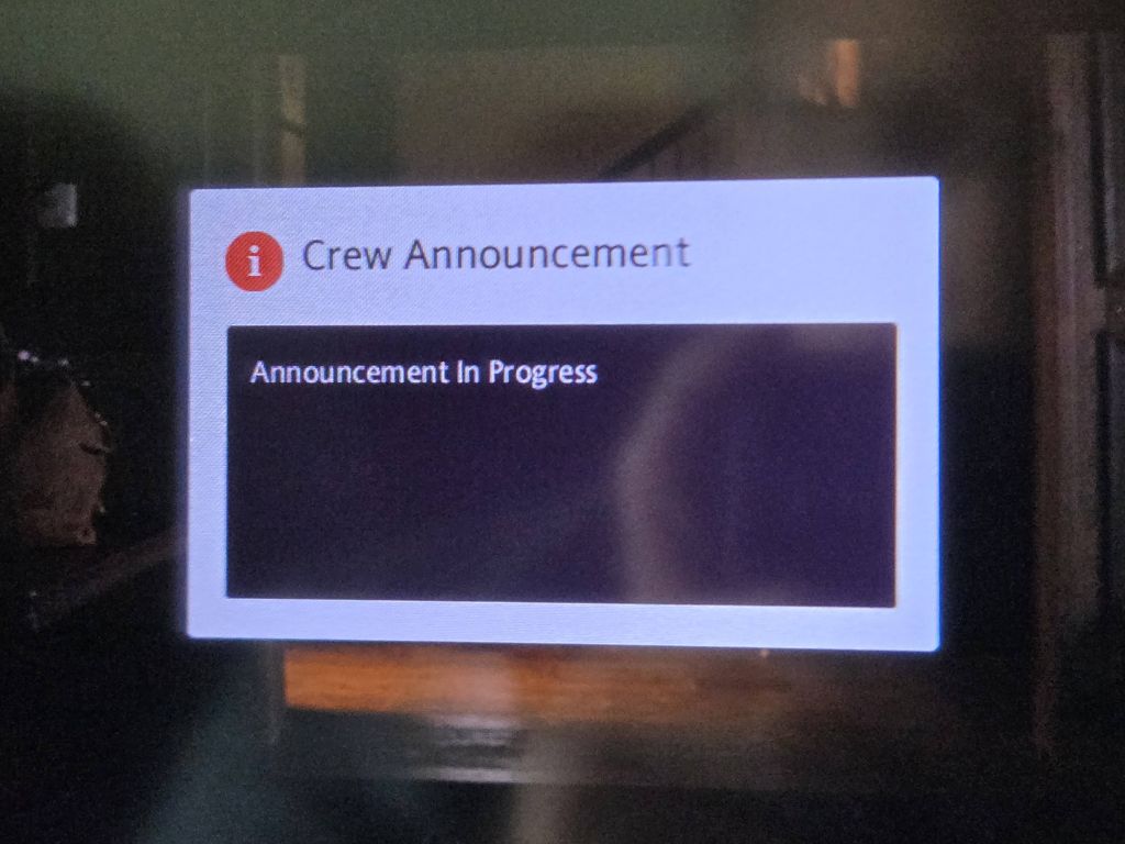 A close up photo of an airplane seatback entertainment screen showing a white popup notification block on a dark background. The block features an orange informational icon, the heading Crew Announcement, and the message Announcement In Progress.