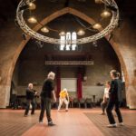 A group of five people practicing movements or rehearsing in the centre of a spacious, high-ceilinged theatre hall built with exposed brick. The room features large arched architectural structures, a tiled floor, gothic-style windows, and a large circular metal truss lighting rig suspended from the ceiling. A projector screen with red curtains stands at the far end of the hall.