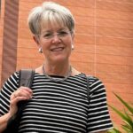 A portrait of Dr Juliette Sterkens smiling outside the Australian Hearing Hub building at Macquarie University. She is a woman with short, grey hair, wearing thin-rimmed glasses, a black and white horizontally striped t-shirt, a silver necklace, and earrings. She has a black backpack strap over her right shoulder. In the background, the large brick wall of the university building features the teal logo and lettering for the Australian Hearing Hub alongside the Macquarie University crest and name.