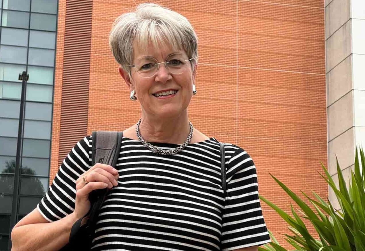 A portrait of Dr Juliette Sterkens smiling outside the Australian Hearing Hub building at Macquarie University. She is a woman with short, grey hair, wearing thin-rimmed glasses, a black and white horizontally striped t-shirt, a silver necklace, and earrings. She has a black backpack strap over her right shoulder. In the background, the large brick wall of the university building features the teal logo and lettering for the Australian Hearing Hub alongside the Macquarie University crest and name.